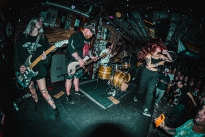 A band performs on stage in a dimly lit venue. The guitarist, bassist, drummer, and vocalist are engaged with their instruments and the energetic crowd in front of them.