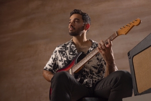 A man with a beard playing an electric guitar while sitting down, next to an amplifier. The background is plain and brown.