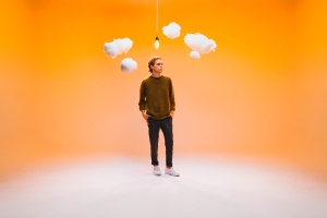 A person stands in a room with an orange background, white fluffy clouds, and a hanging light bulb. They are wearing a green sweater and black pants.