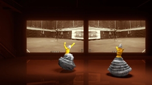 Two dancers in long, striped skirts and yellow tops perform in a dark room in front of two large screens displaying identical sepia-toned scenes.