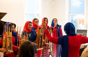 A music conductor directs a group of women in colorful attire, some holding bamboo percussion instruments, during a performance in a well-lit room.