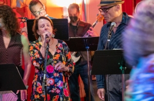 A group of people perform on stage. A woman in a floral dress sings into a microphone while others play instruments and sing in the background.