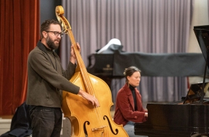 A man plays a double bass while a woman plays the piano in a room with red and grey curtains.