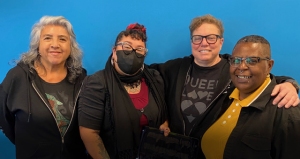 Four people stand together in front of a blue background. One person wears a black mask, and another has their arm around a friend. They all smile at the camera.