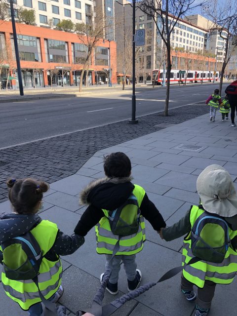 穿著反光背心和背包的孩子們手牽手走在人行道上，背景是一條有電車的街道。