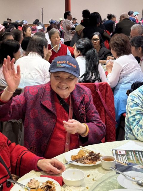 一位戴著「麥法恩競選市長」帽子的年長婦女在擁擠的聚會中微笑著揮手。她坐在一張擺有食物的桌子旁。