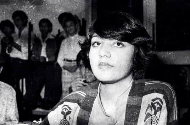 A sepia-toned photograph of a young person with medium-length hair looking at the camera while others in the background appear blurred.