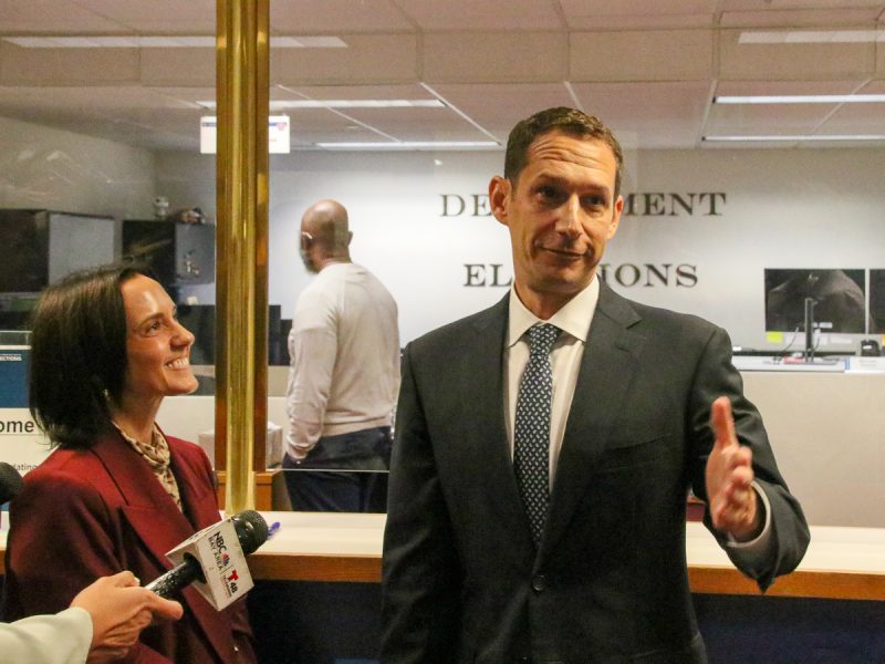 A man in a suit gestures while speaking, with a woman standing beside him. They appear to be in an office environment with a sign reading "DEPARTMENT OF ELECTIONS" visible in the background.