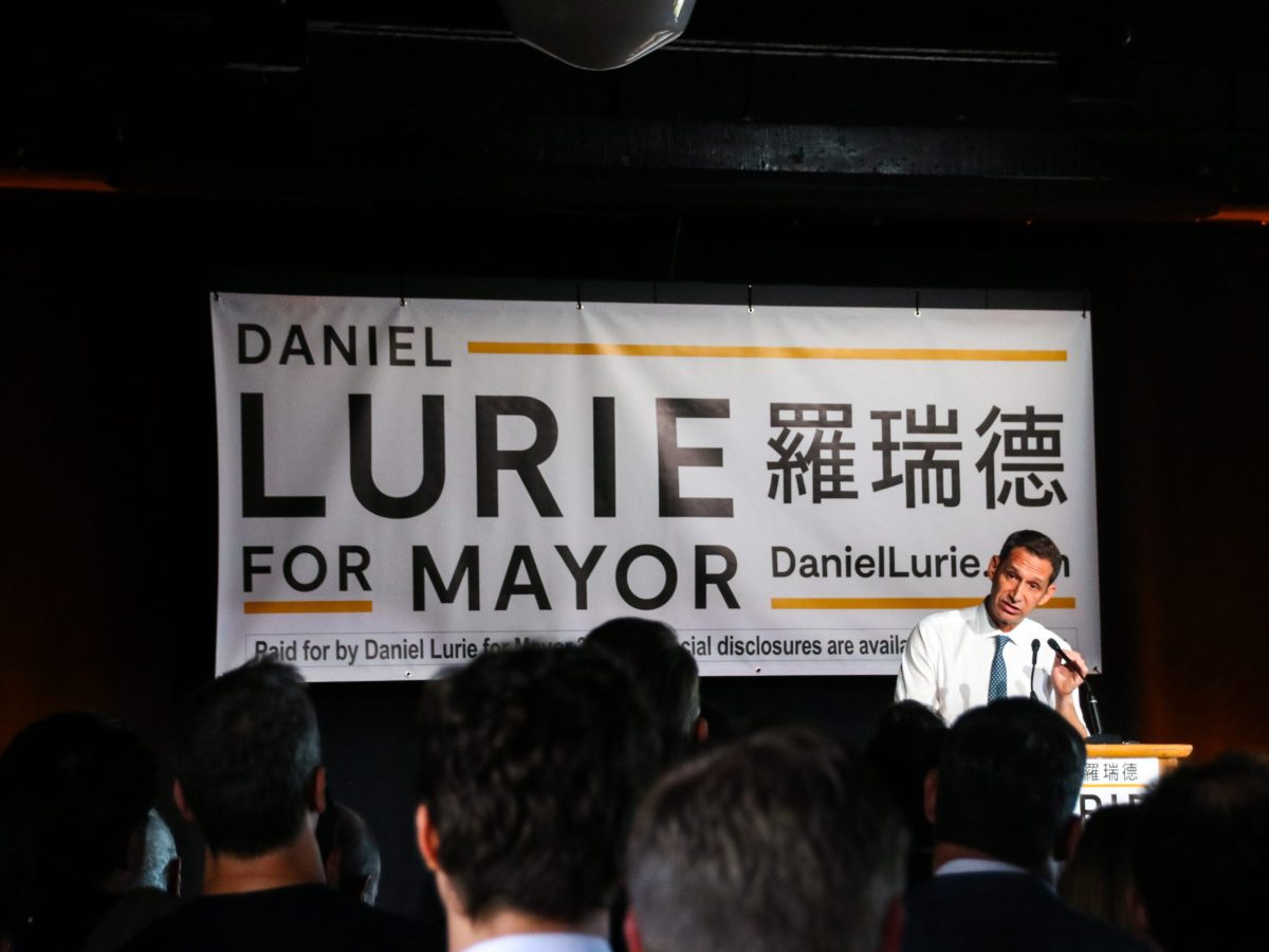 A man speaks at a podium in front of a banner that reads "Daniel Lurie for Mayor" with text in Chinese characters and a website link, addressing a crowd.