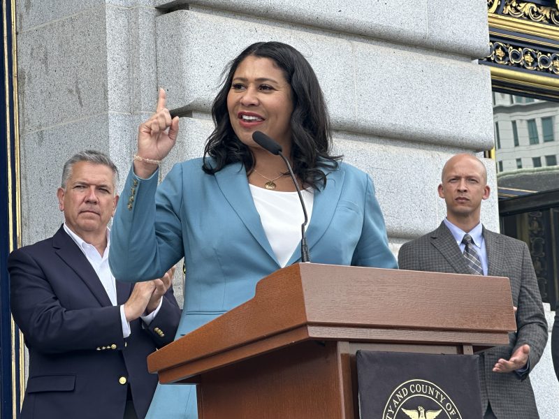 Mayor London Breed in a blue blazer speaks at a podium outside a building, with two men standing behind her.