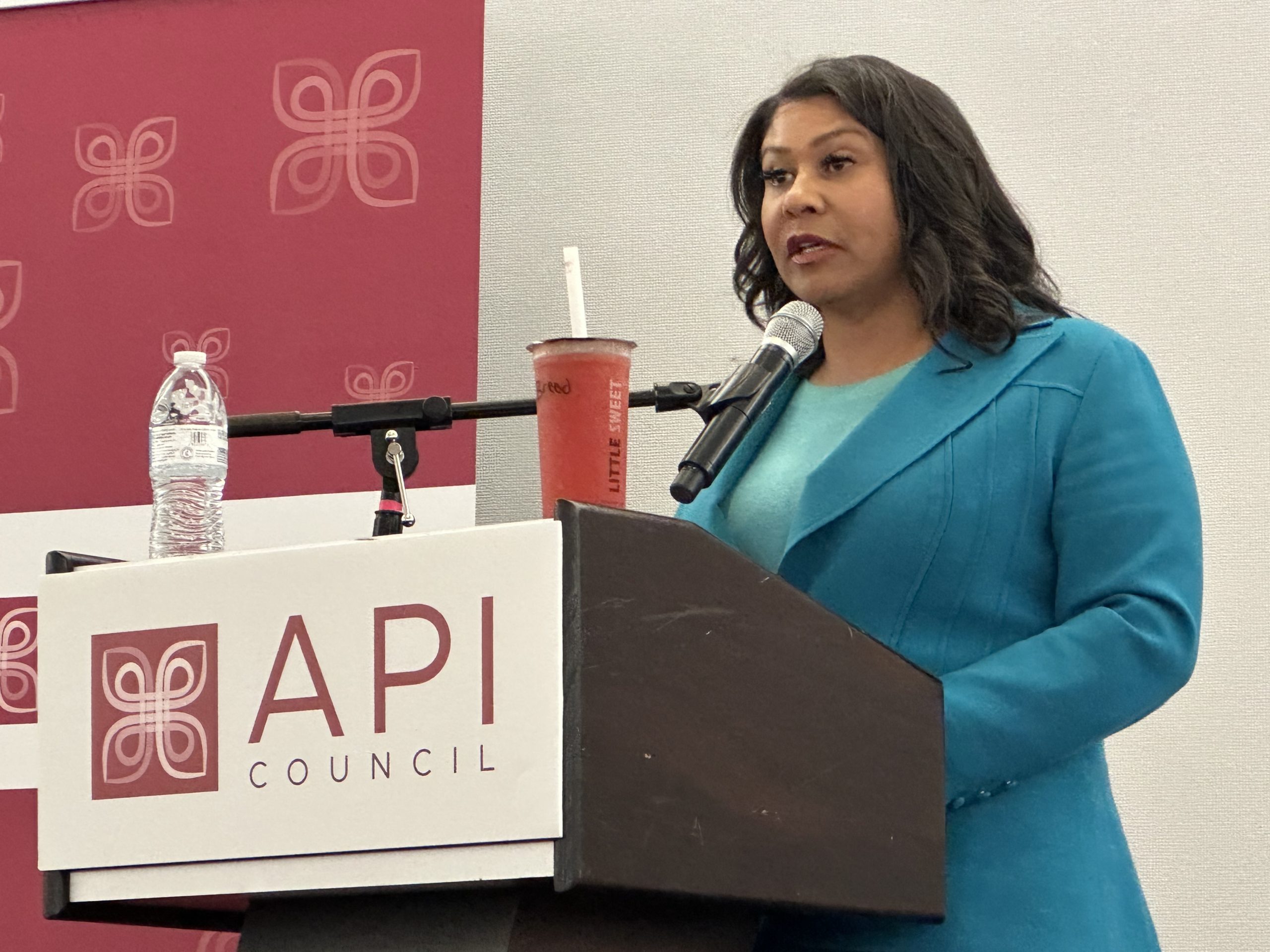 San Francisco Mayor London Breed stands at a podium labeled "API Council," speaking into a microphone in Chinatown. A water bottle and a drink are placed on the podium.