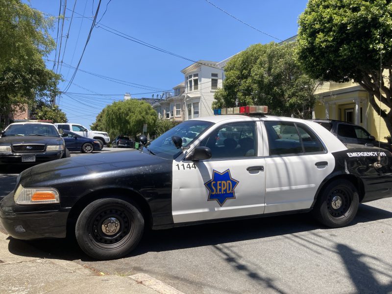 A black and white police car is parked on a residential Powell Street, marked with the text "S.F.P.D." on the side and a visible emergency light on the roof.