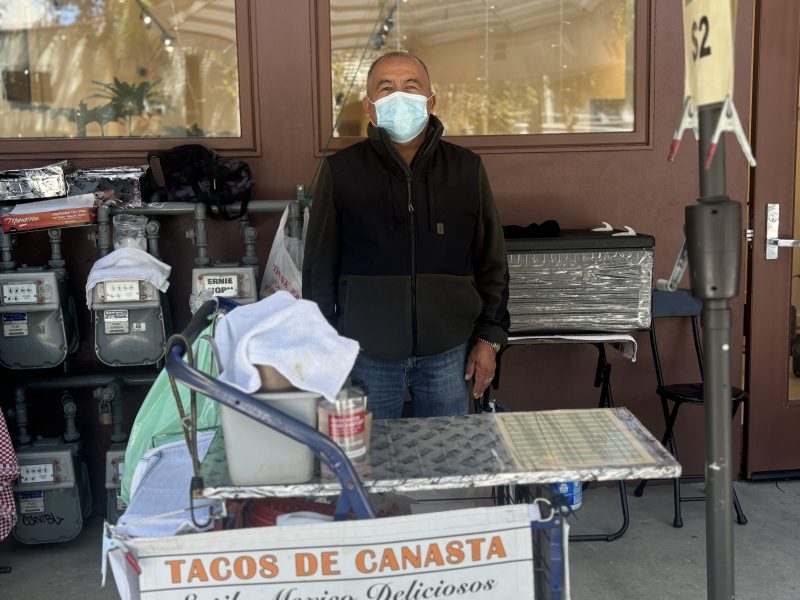 A man wearing a mask stands behind a street food cart labeled "Tacos de Canasta" in front of a building, with gas meters and various items in the background.
