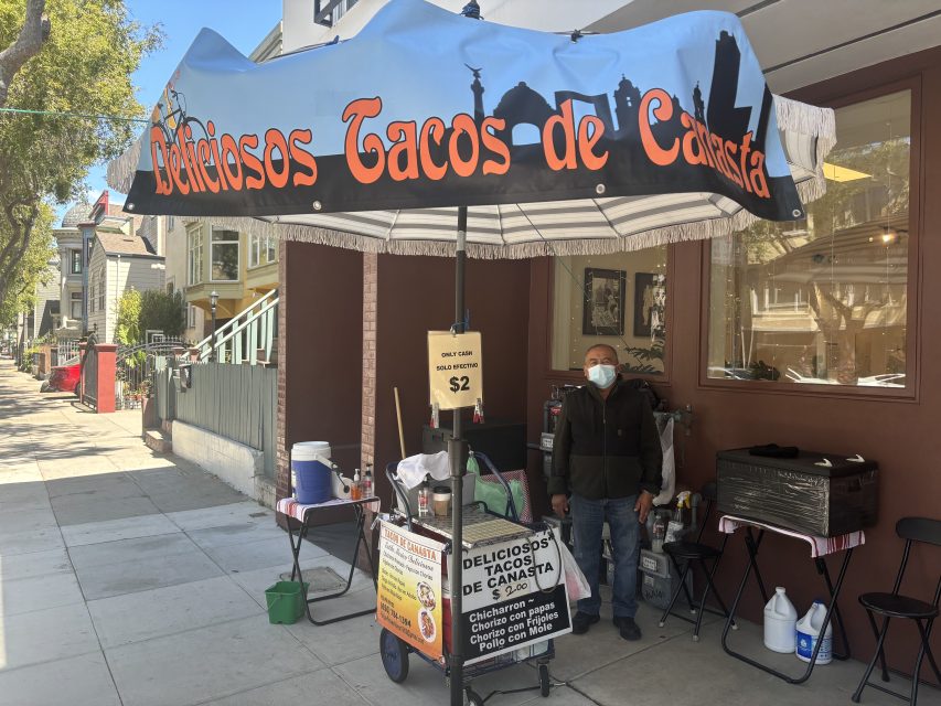 A person stands behind a taco stand on the sidewalk, selling "Deliciosos Tacos de Canasta" for $2 each and offering a variety of taco options. An umbrella shades the stand.