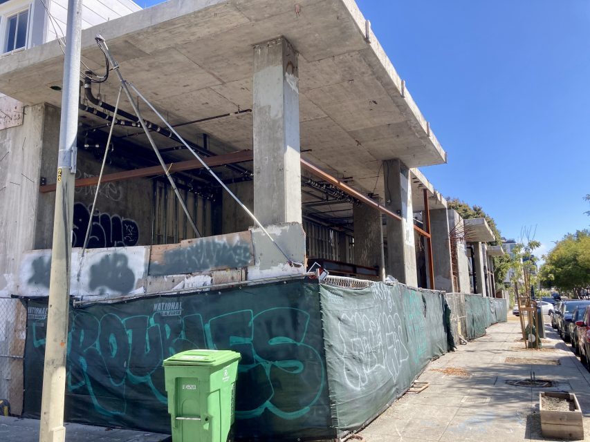A partially constructed concrete building with graffiti on the fencing and a green trash bin nearby, under clear blue skies.