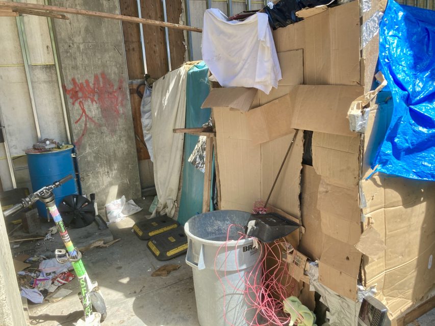 A makeshift shelter constructed with cardboard boxes, tarps, and various items including a garbage bin, scooter, and tools. Clothing is draped over the structure.