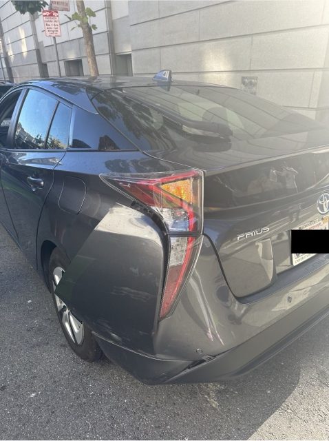 A dark grey Toyota Prius is parked on a street next to a building. There is noticeable damage to the rear left corner and scratched paint. A partially blacked-out license plate is visible.