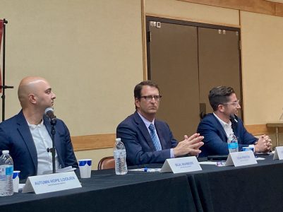Bilal Mahmood, Dean Preston and Scotty Jacobs are seated behind a panel table with name placards, microphones, and water bottles in front of them. The person in the middle is clasping their hands while listening.