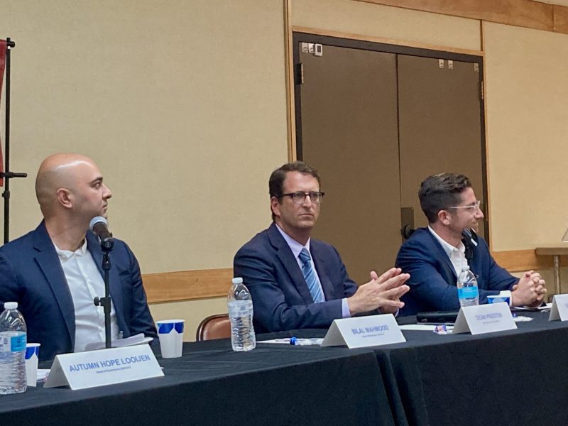 Three men are seated at a panel discussion table with microphones and water bottles. Name cards are visible in front of each panelist. The middle man is clapping while the others are looking to their left.