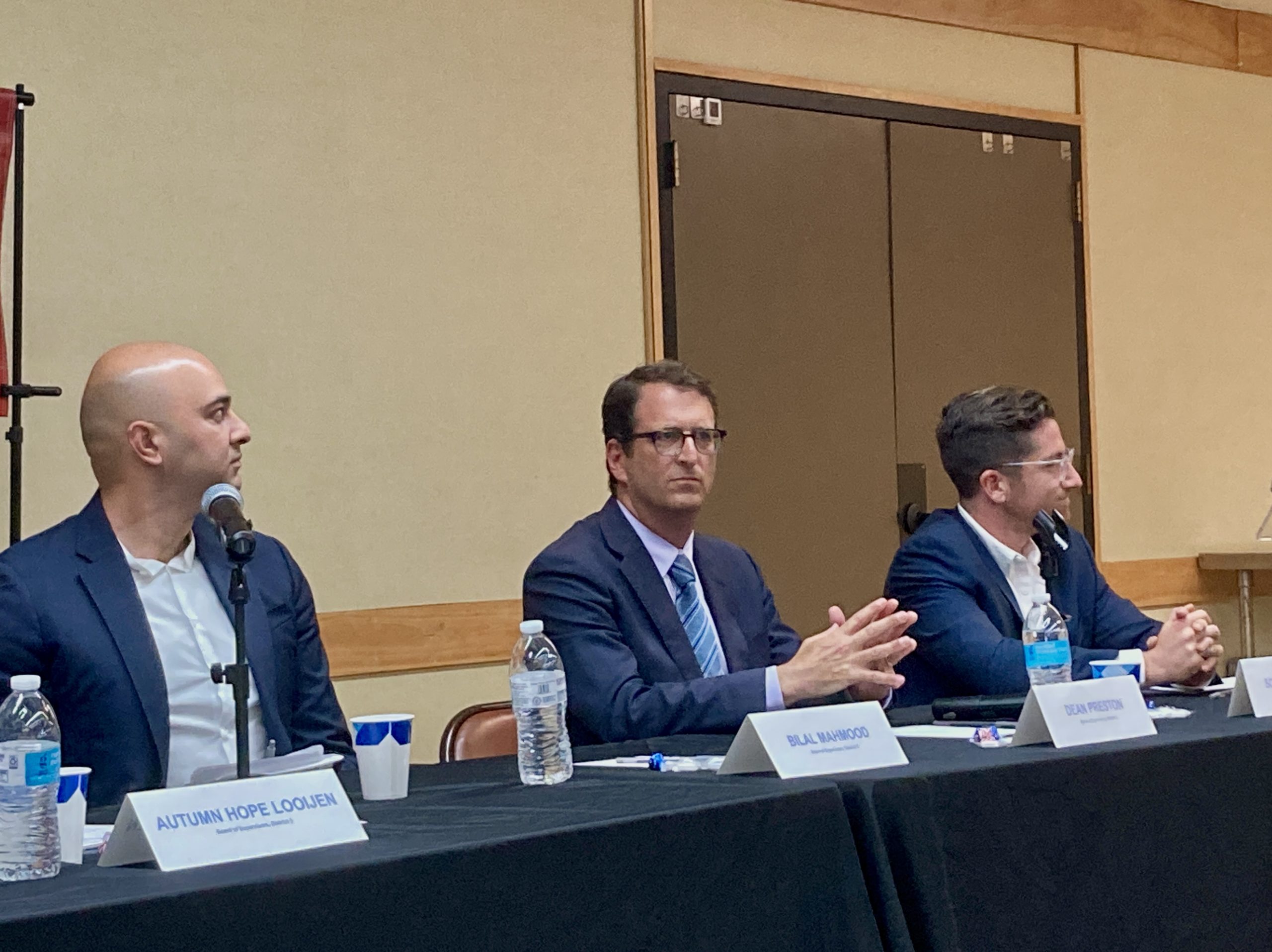 Bilal Mahmood, Dean Preston and Scotty Jacobs are seated behind a panel table with name placards, microphones, and water bottles in front of them. The person in the middle is clasping their hands while listening.