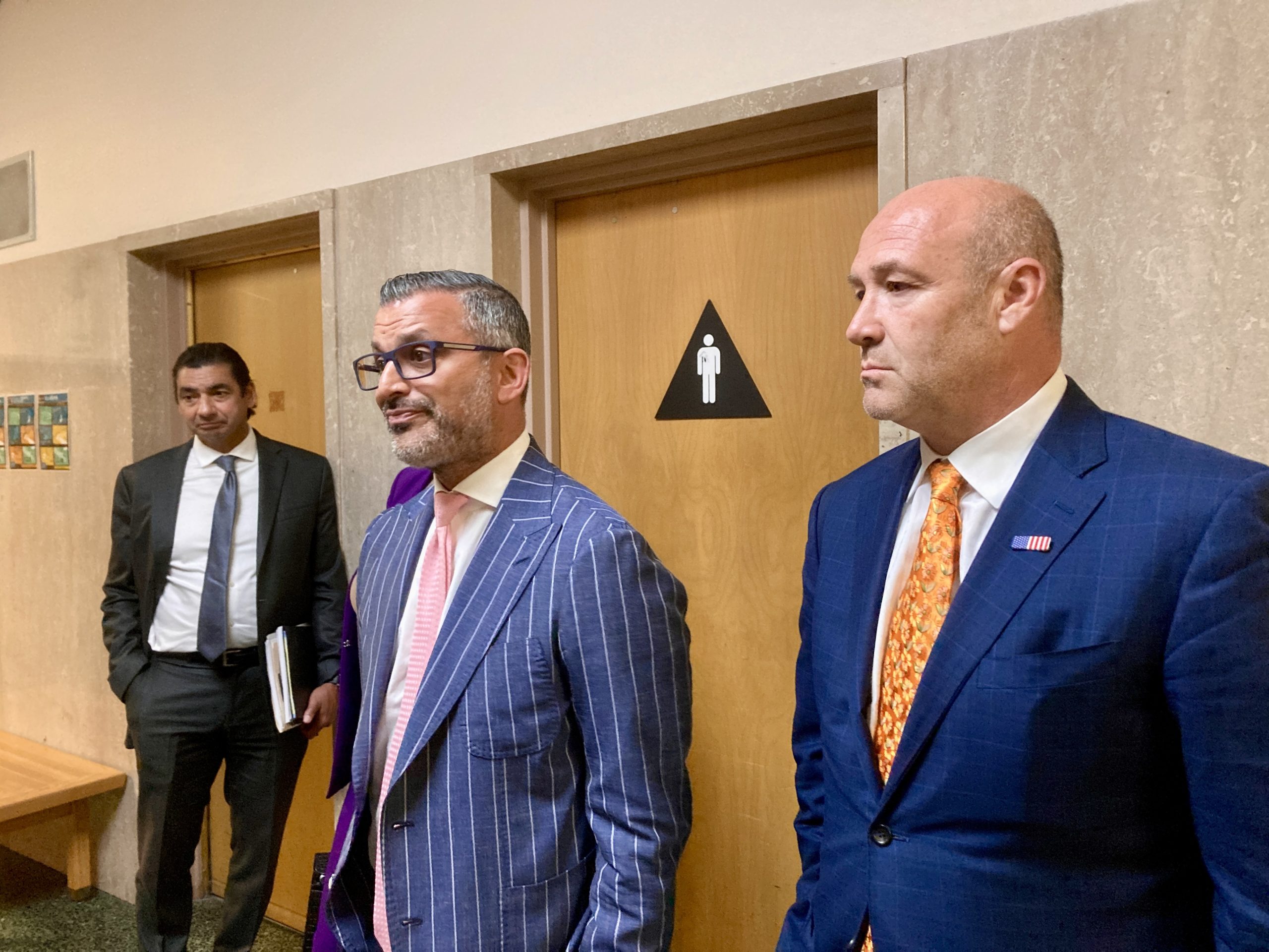 Three men in suits stand in a hallway. Two of them are positioned near a door marked with a restroom symbol. One man holds a folder and another man stands in the background.