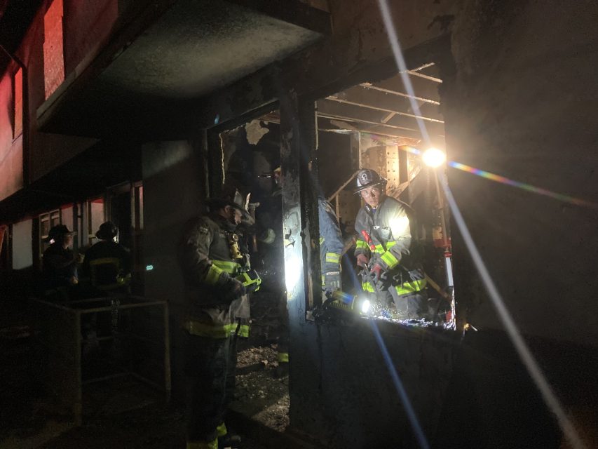 Firefighters in gear assess damage through a window of a charred, dimly lit building in Potrero Hill.