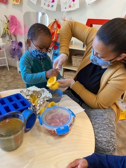 一個孩子和一個成年人，都戴著口罩，坐在桌旁擠檸檬汁到碗裡，周圍擺放著水果和容器。他們身後的牆上展示著藝術作品。