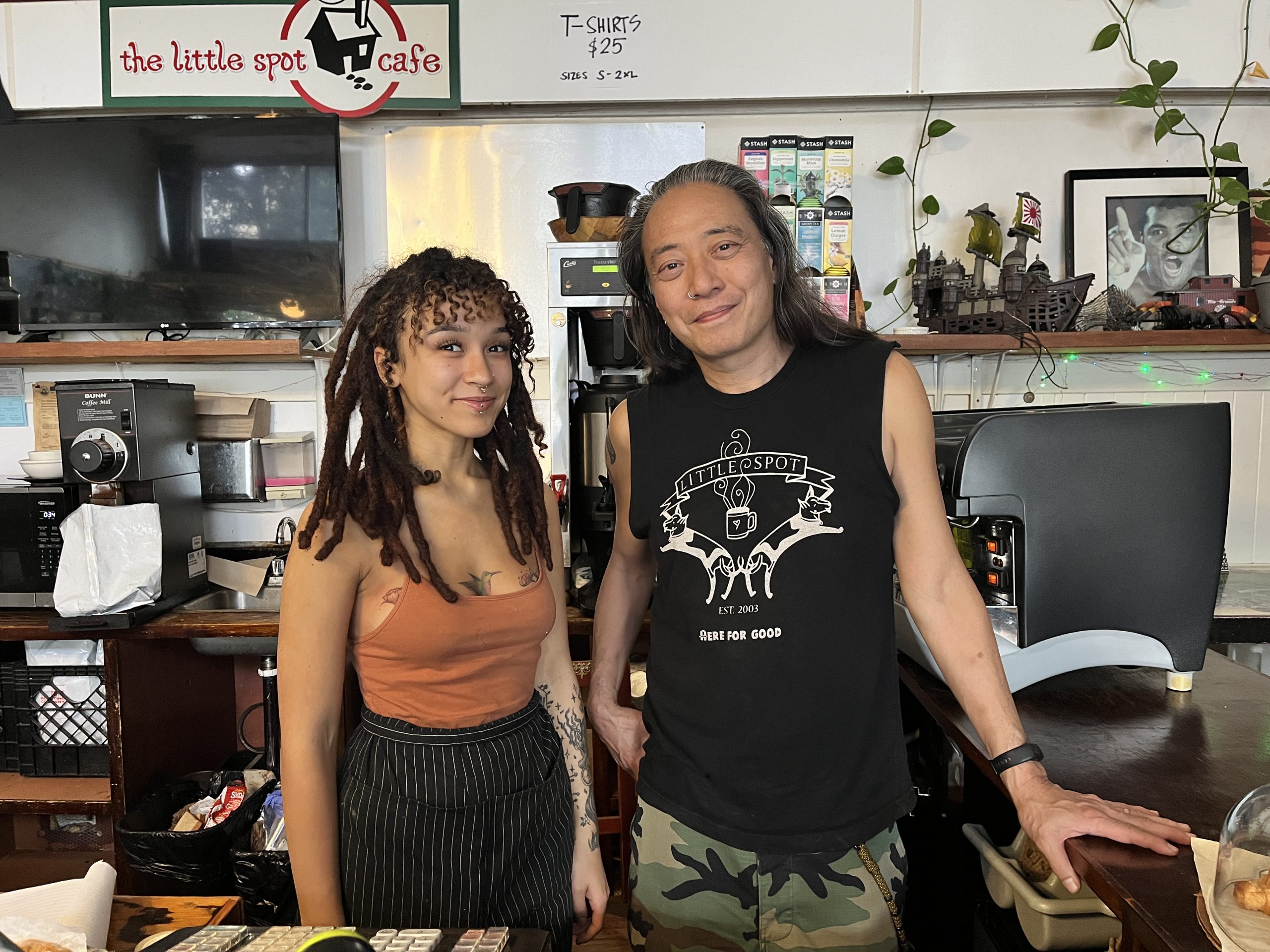 Two people stand behind the counter of The Little Spot Cafe. One has long dreadlocks and wears an orange top, while the other has long hair and wears a sleeveless black shirt with camo pants.