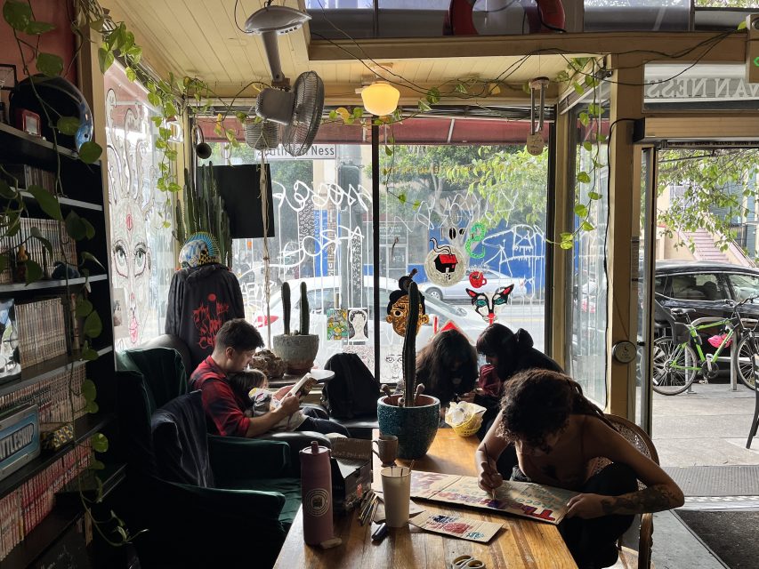 A group of people are seated at a table inside a cozy café, engaged in various activities. The space has green plants, large windows, and colorful decorations, with bicycles visible outside.
