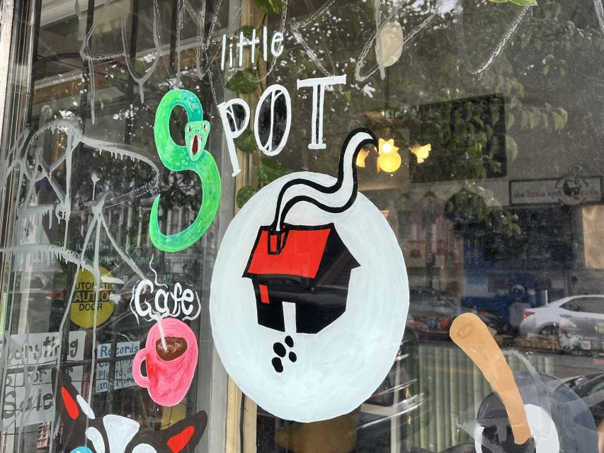 A cafe window with a painted sign that reads "little SPOT Cafe" featuring a green 'S' and a circular logo of a black house with a red roof and chimney smoke.