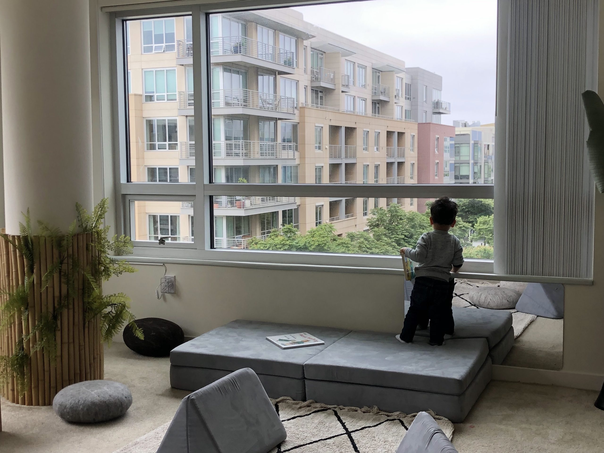一個孩子站在大窗戶旁的坐墊上，手持一本書，望向外面現代公寓樓和綠地的景色。室內擺放著植物和散落的坐墊。