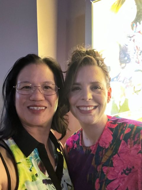 Two women posing for a photo together, both smiling. The woman on the left is wearing glasses and a sleeveless patterned top, while the woman on the right is wearing a vibrant floral dress.