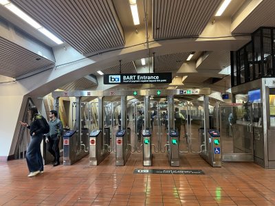 BART station entrance showing multiple turnstiles, with passengers entering and exiting. A sign indicates that BART cards or Metro passes are required for entry.