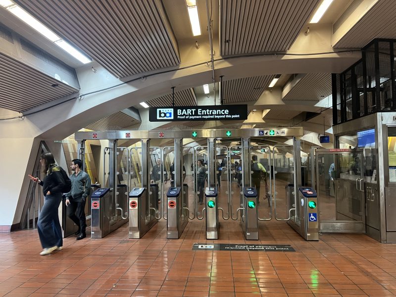 BART station entrance showing multiple turnstiles, with passengers entering and exiting. A sign indicates that BART cards or Metro passes are required for entry.