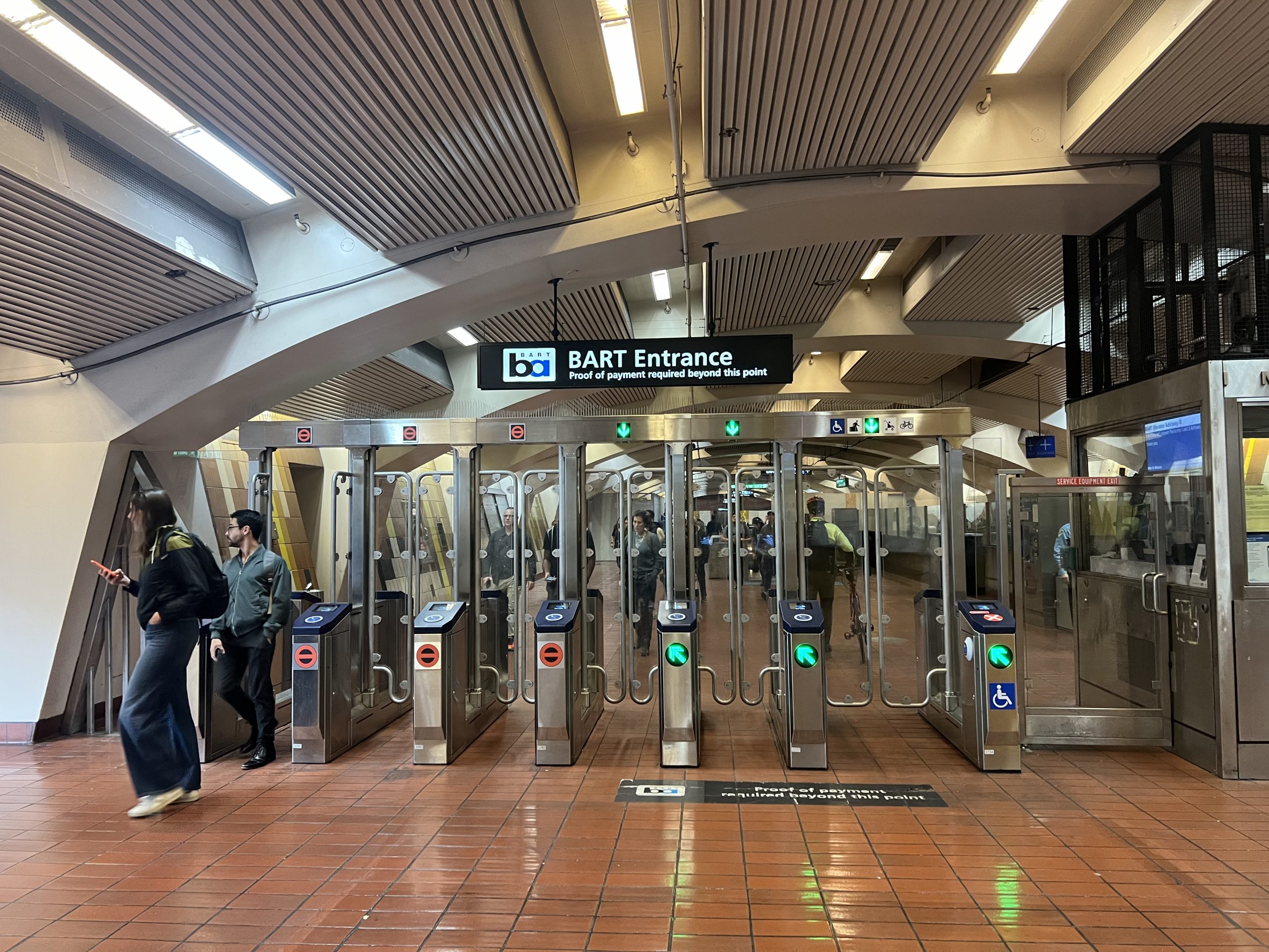 BART station entrance showing multiple turnstiles, with passengers entering and exiting. A sign indicates that BART cards or Metro passes are required for entry.