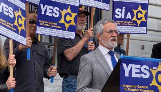 A group of people hold "YES on C" signs advocating for an Inspector General to combat corruption, as a man in a gray suit speaks at a podium.