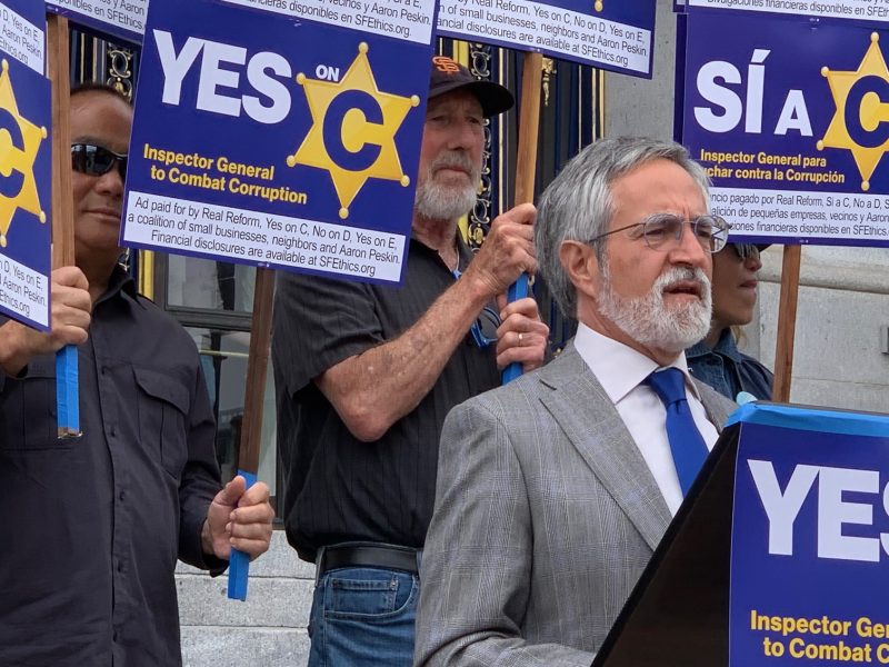 A group of people hold "YES on C" signs advocating for an Inspector General to combat corruption, as a man in a gray suit speaks at a podium.