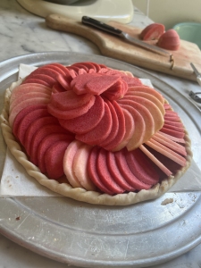 Pie with neatly arranged slices of red and pink apples on a baking sheet, with a wooden cutting board and knife in the background.