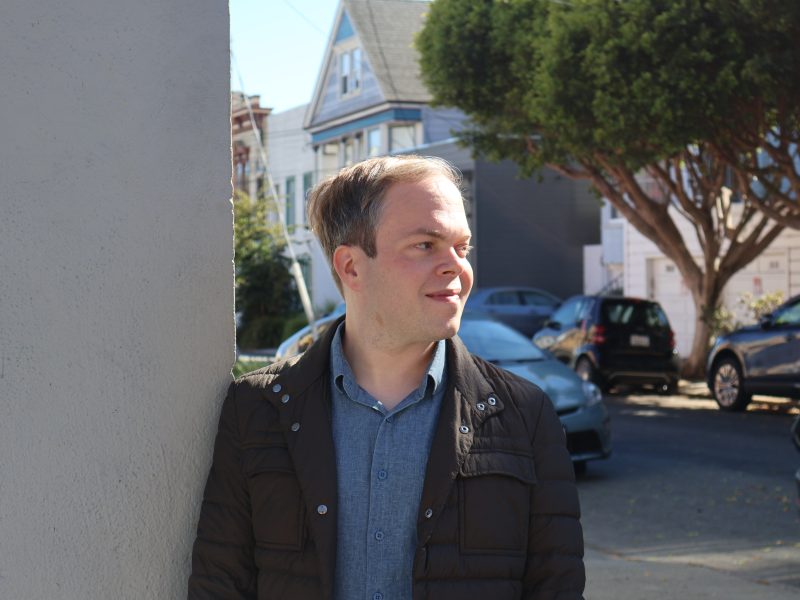 A man with short hair leans against a wall outdoors in a residential neighborhood, looking to the right. Cars are parked along the street, and houses and trees are in the background.