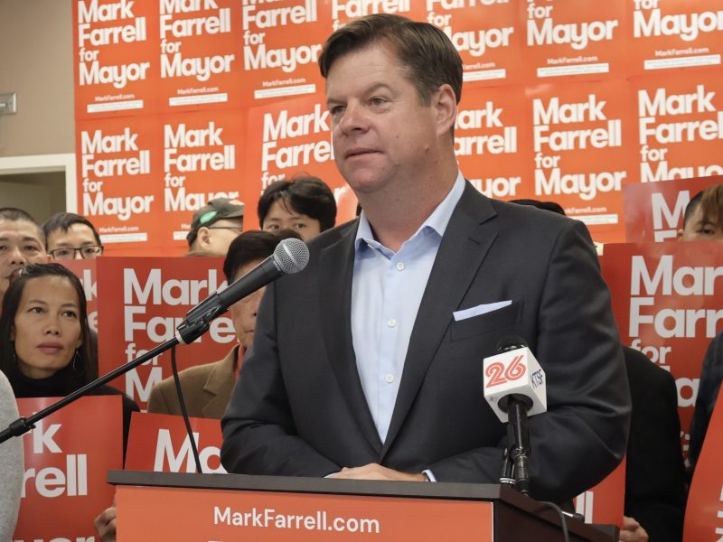 A man in a suit speaks into a microphone at a podium, surrounded by people holding "Mark Farrell for Mayor" signs.