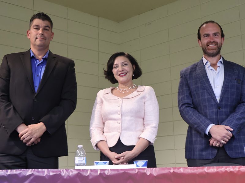 Three people stand behind a table with water bottles and cups. The wall behind them is white and tiled, and they appear to be at an indoor event or meeting.