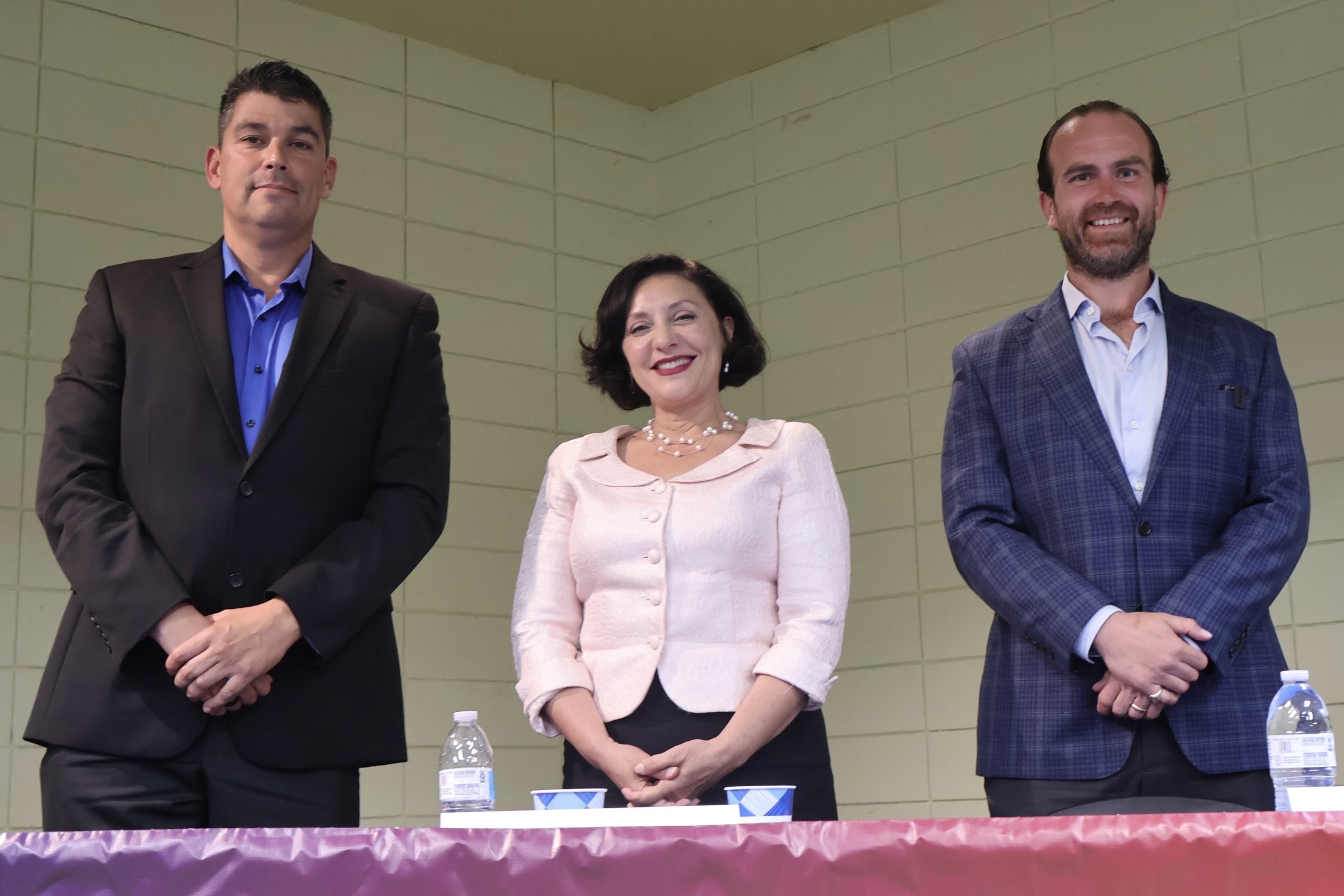 三個人站在一張擺放著水瓶和杯子的桌子後面。他們身後的牆壁是白色的瓷磚，看起來他們正在參加一個室內活動或會議。