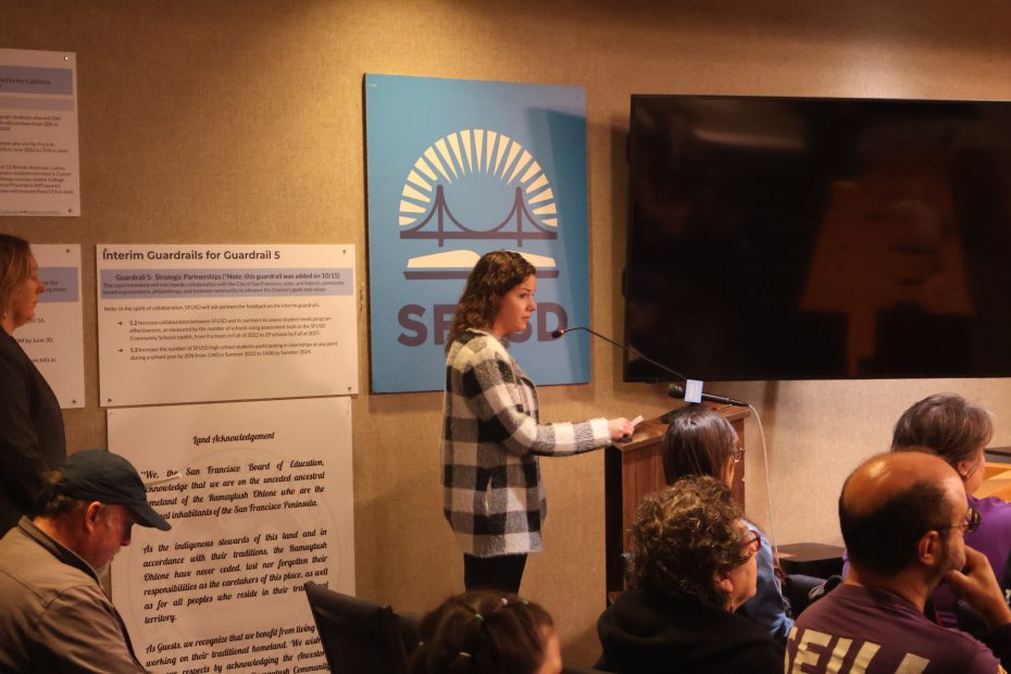 A woman speaks at a podium in a room with seated attendees. She stands next to a large screen and a wall with informational posters about SFUSD.