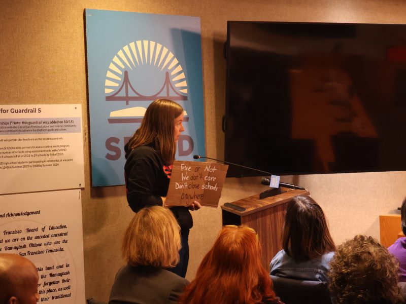 A person speaks at a podium in a room with several seated attendees. They hold a cardboard sign with handwritten text. A large screen and an SFUSD sign with a bridge graphic are visible behind them.