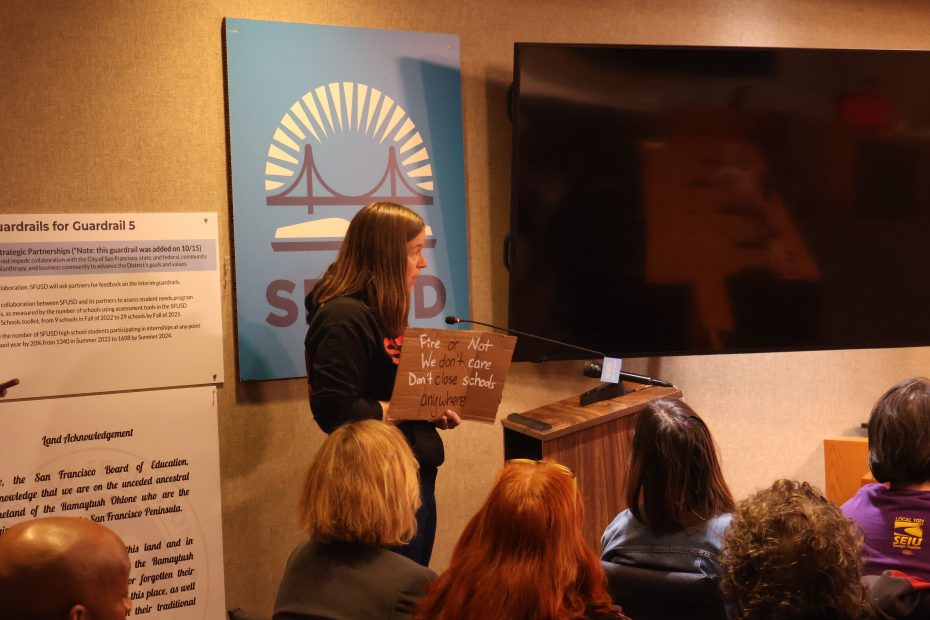 A person speaks at a podium in a room with several seated attendees. They hold a cardboard sign with handwritten text. A large screen and a sign with a bridge graphic are visible behind them.