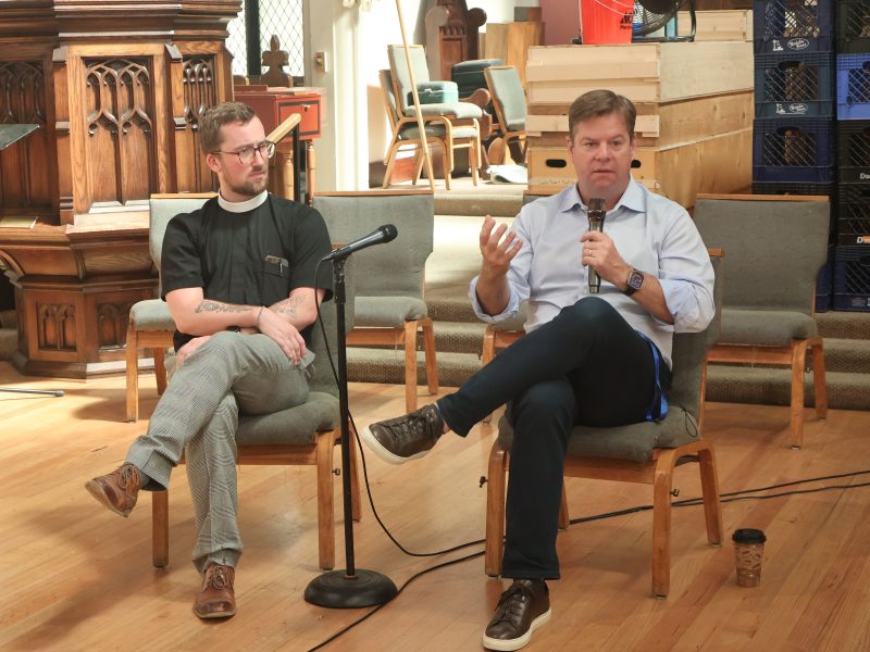 Two men are seated on chairs, engaged in a discussion with microphones, inside a room furnished with wooden furniture and chairs. One man is gesturing with his hand while speaking.