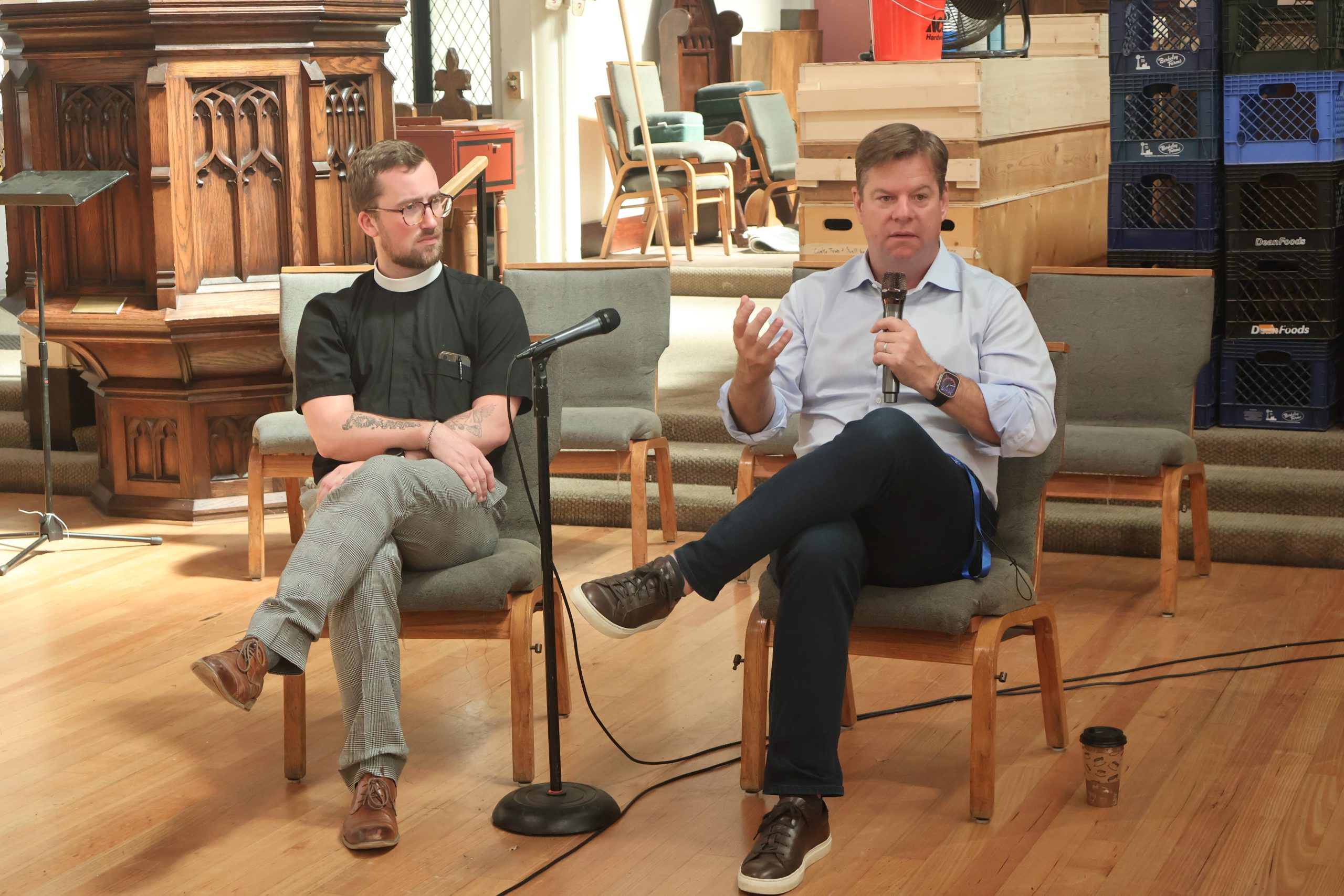兩名男子坐在椅子上，手持麥克風進行討論，他們身處一個擺放著木製家具和椅子的房間內。其中一名男子在說話時正用手做著手勢。