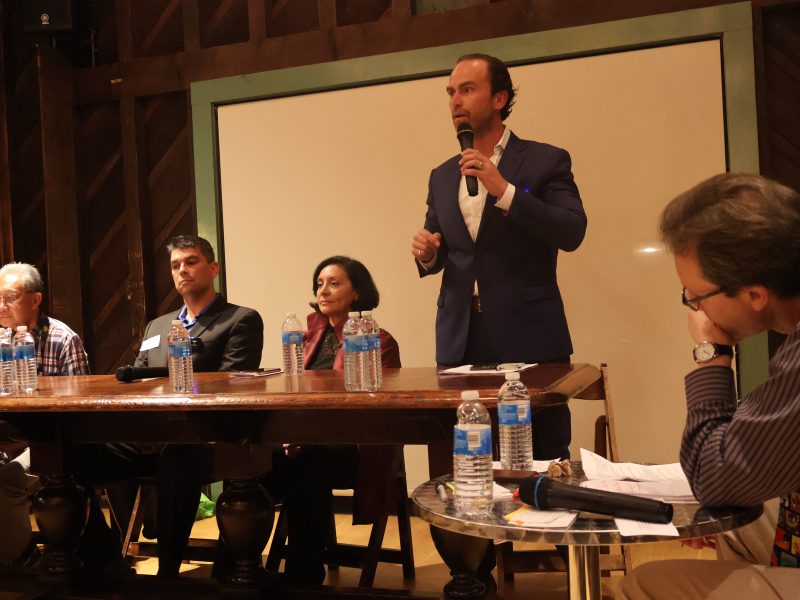 A panel discussion with five speakers seated at a table in front of a whiteboard. One speaker is standing, speaking into a microphone. Water bottles and microphones are on the table.