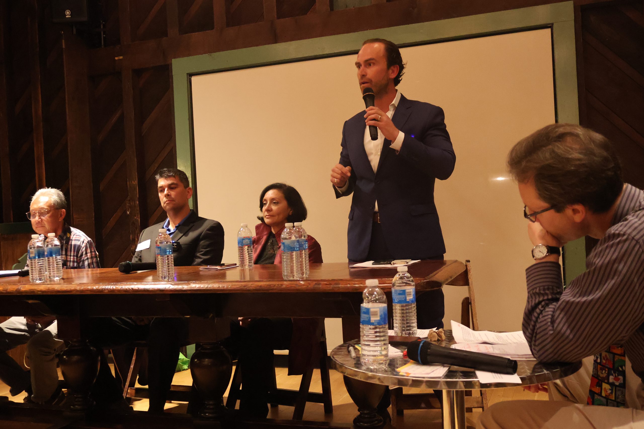 A panel discussion with five speakers seated at a table in front of a whiteboard. One speaker is standing, speaking into a microphone. Water bottles and microphones are on the table.