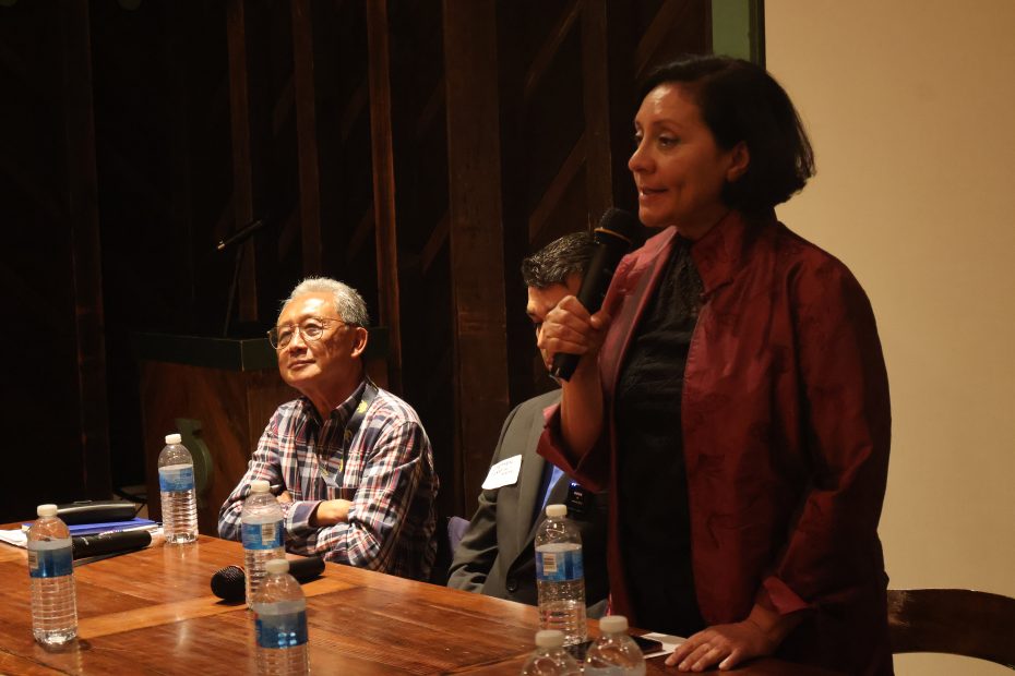 Three individuals are seated at a table with water bottles. One person standing and speaking into a microphone.